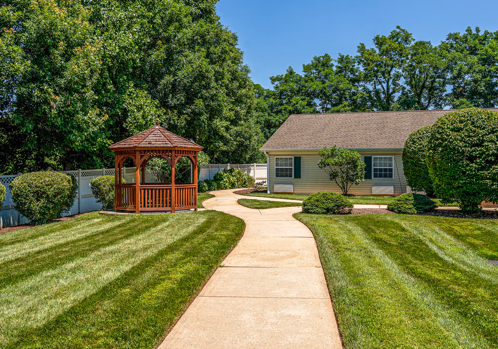 A landscaped outdoor courtyard with a wooden gazebo, curving sidewalk, manicured lawns and a small single-story building.