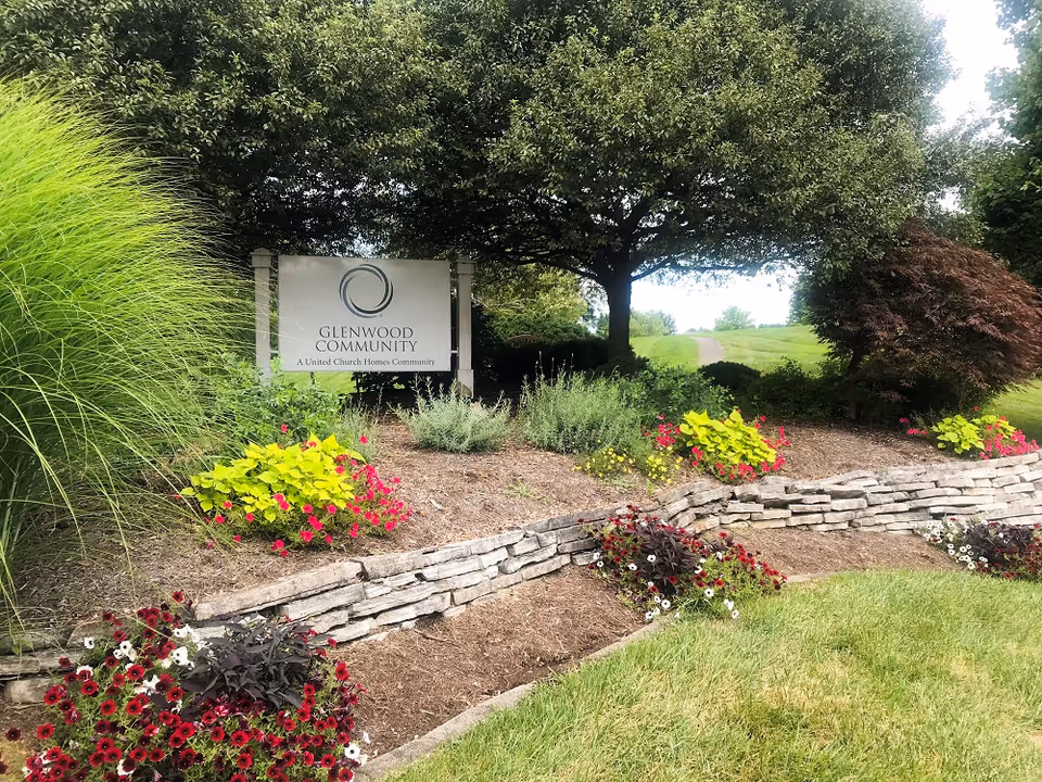 A landscaped garden area with colorful flowers and greenery surrounding a sign that reads 'Glenwood Community, A United Church Homes Community' under a large tree with a grassy path in the background.
