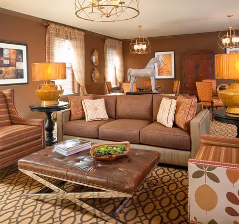 A cozy living room with a brown sofa adorned with patterned pillows, two striped armchairs, and a large leather ottoman coffee table with a decorative bowl and books on it. The room features warm brown walls, patterned carpet, two table lamps with yellow bases, and three hanging light fixtures. There are framed artworks and decorative plates on the walls, and a wooden horse sculpture on a table in the background.