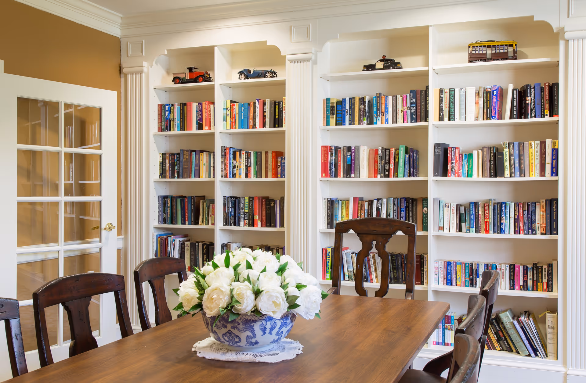 A cozy reading room with white built-in bookshelves filled with books and model cars on top. In front of the shelves is a wooden table with a blue and white vase holding a bouquet of white flowers. Several wooden chairs surround the table. A glass-paneled door is visible on the left side of the room.