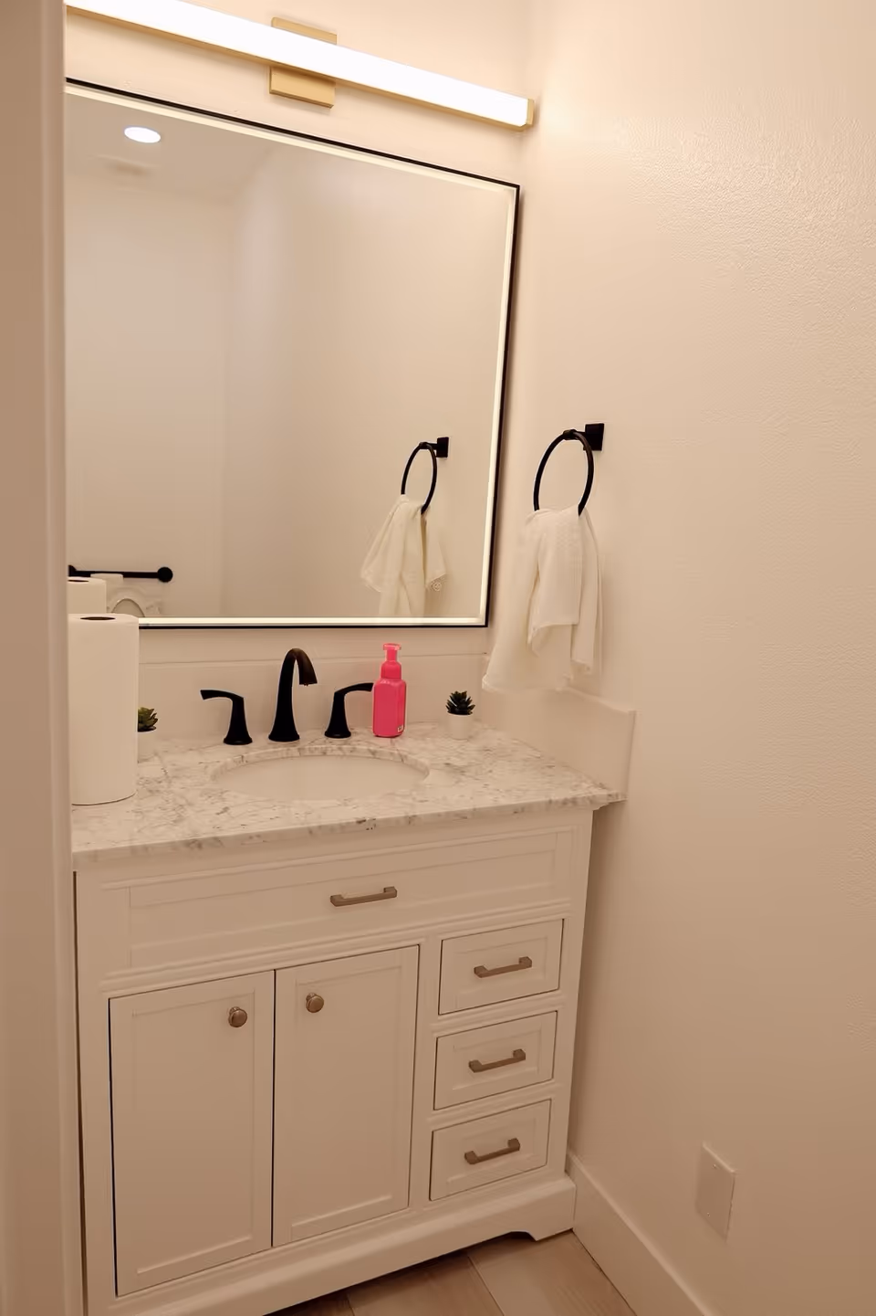 A clean bathroom vanity with a white marble countertop, black faucet fixtures, a large rectangular mirror with a light above it, a white towel hanging on a black towel ring, a pink soap dispenser, two small potted plants, and a roll of paper towels.