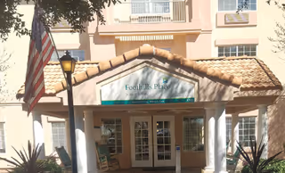 Entrance of a senior living facility named Foothills Place with a tiled roof, white columns, an American flag on a pole, and a lamp post in front. The building has beige walls and multiple windows.