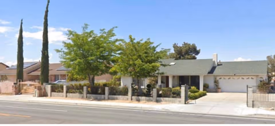 Single-story residential building with a green roof, a front yard with trees and shrubs, and a driveway leading to a two-car garage. The property is enclosed by a low fence with stone pillars, and there are tall cypress trees on the left side. The sky is clear with a few clouds.