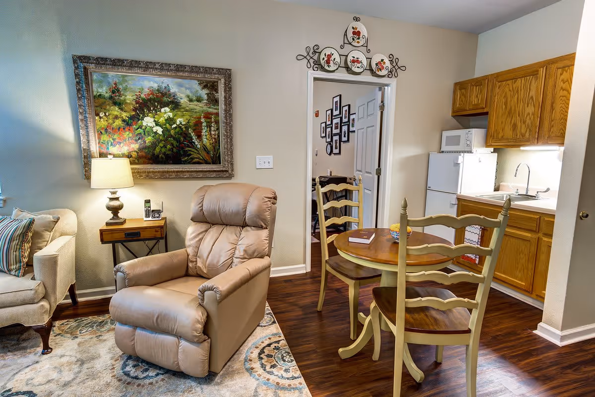 A cozy living area with a beige recliner chair, a small wooden side table with a lamp and phone, and a beige sofa with striped pillows. Adjacent to the living area is a small dining table with two chairs and a kitchen area with wooden cabinets, a white refrigerator, microwave, and sink. A framed floral painting hangs on the wall above the recliner, and decorative plates are displayed above the doorway leading to another room.