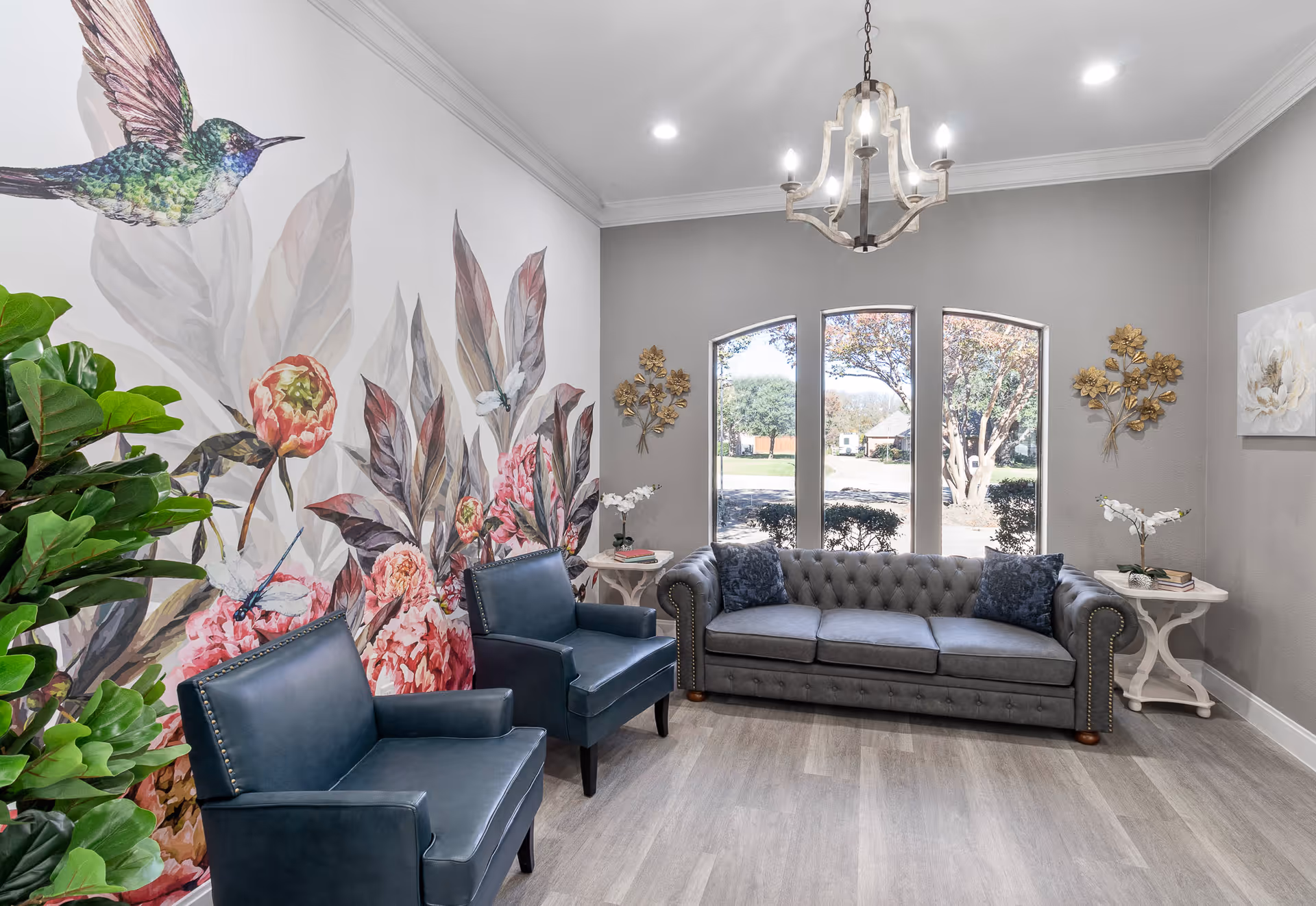 A bright and cozy living room with a large floral and hummingbird mural on one wall. The room features two dark blue armchairs and a gray tufted sofa with two dark blue pillows. There are two small white side tables with decorative items, including flowers and books. Three tall windows let in natural light, and a chandelier hangs from the ceiling. The walls are painted light gray, and the floor has light wood-style flooring.