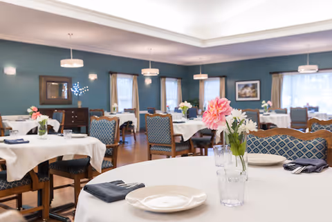 A spacious dining room with multiple tables covered in white tablecloths, each set with plates, glasses, and napkins. The tables have floral centerpieces with pink and white flowers. The room features blue walls, several windows with beige curtains, wooden chairs with blue patterned upholstery, and soft ceiling lighting fixtures.