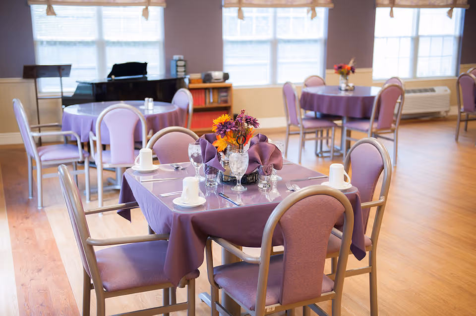 A dining room with round and square tables covered with purple tablecloths and set with cups, glasses, and silverware. Each table has a centerpiece with colorful flowers. The room has wooden flooring, large windows with blinds, and a black piano in the background.