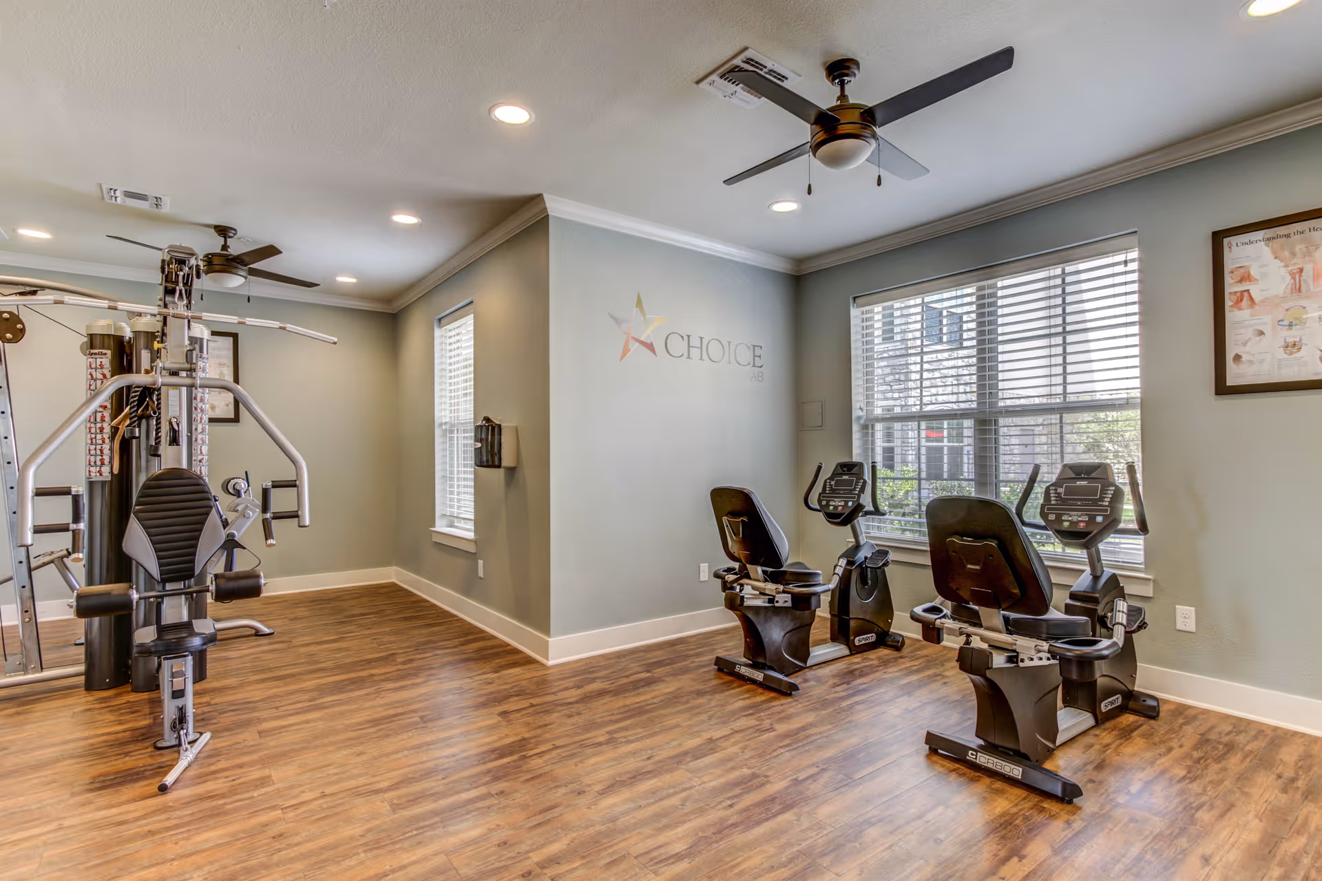 A senior living facility fitness room with exercise equipment including a multi-station weight machine and two recumbent exercise bikes. The room has wood flooring, light green walls, two windows with blinds, ceiling fans, and recessed lighting. There is a framed anatomical poster on the wall and a logo that reads 'CHOICE' on one wall.