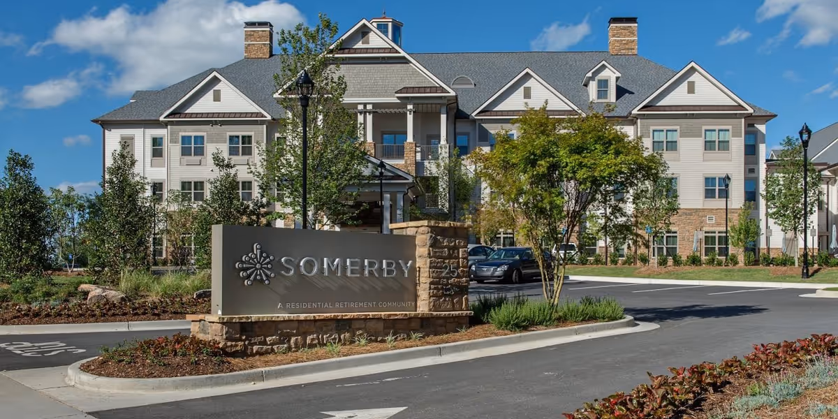 Exterior front view of Somerby Sandy Springs Senior Living facility showing a large multi-story building with white and gray siding, stone accents, multiple windows, and a peaked roof under a blue sky with some clouds. In front of the building is a landscaped area with trees, shrubs, and a stone sign that reads 'SOMERBY A RESIDENTIAL RETIREMENT COMMUNITY'. There is a paved driveway and parking area with a few cars visible.
