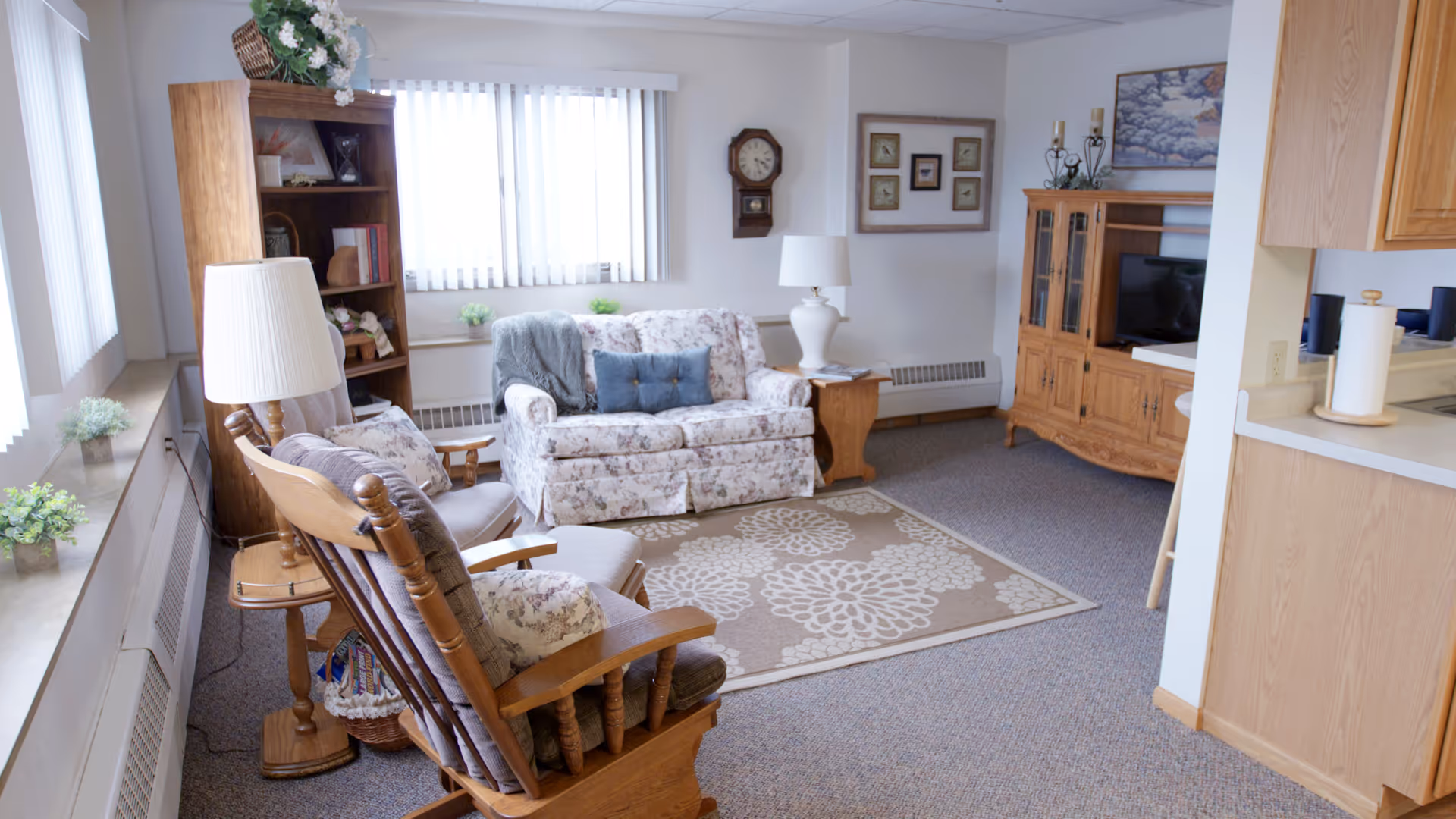 A cozy living room with a floral patterned loveseat, two wooden armchairs with cushions, a wooden bookshelf filled with books and decorative items, a wooden TV cabinet with a television, a side table with a lamp, a wall clock, framed pictures on the wall, and a beige rug with floral patterns on the carpeted floor. The room has large windows with vertical blinds allowing natural light to enter.
