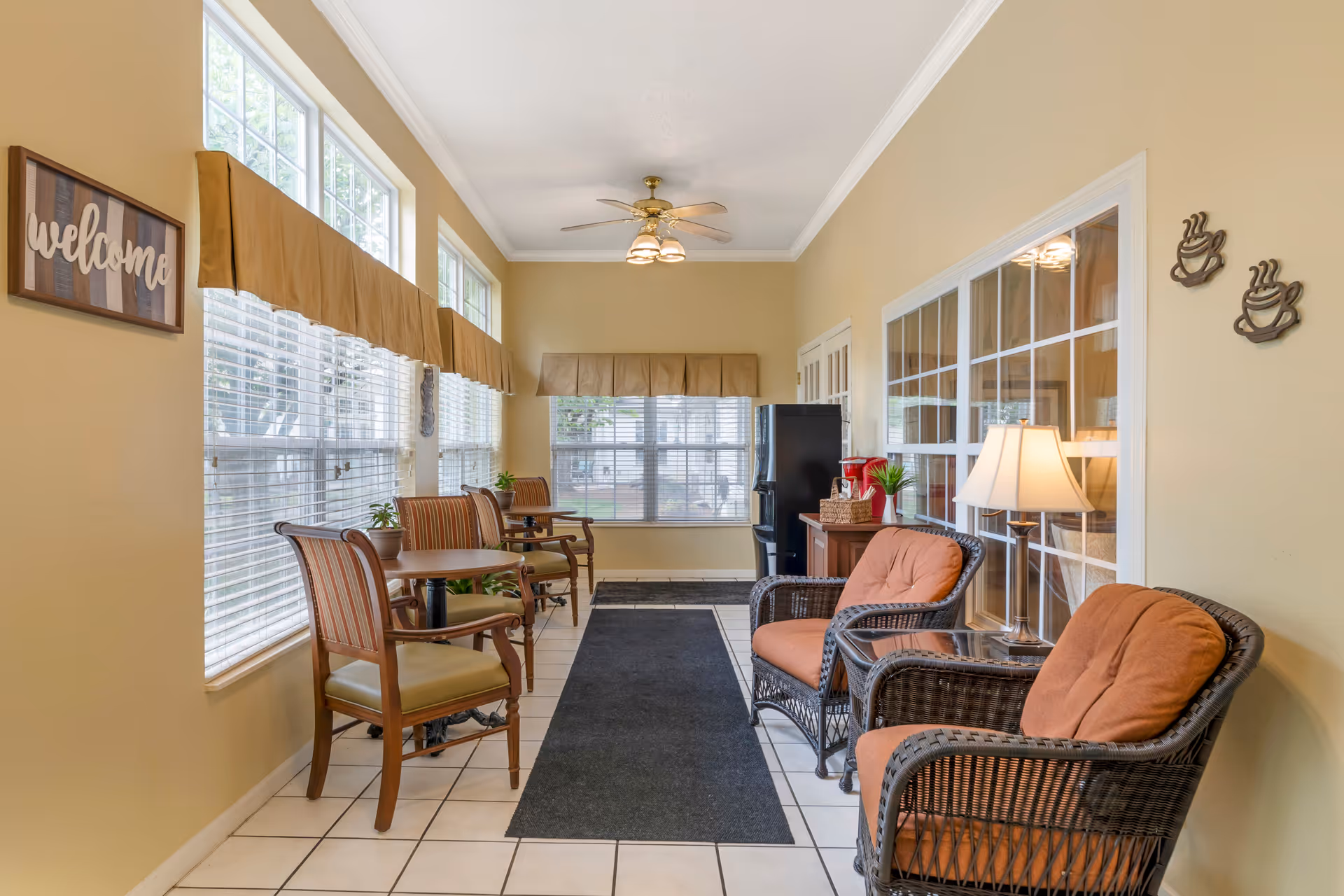 A bright and welcoming sitting area with large windows on the left side, letting in natural light. The room features a row of wooden chairs with cushions and small round tables along the window wall. On the right side, there are two wicker armchairs with orange cushions, a side table with a lamp, and a small cabinet with a coffee maker. The walls are painted light yellow, and there is a ceiling fan with lights. A 'welcome' sign is hung on the left wall near the windows.