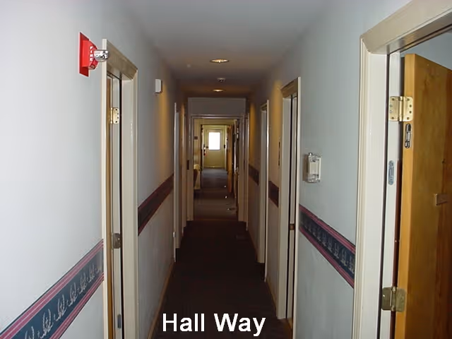 A narrow hallway in a facility with white walls, a dark carpeted floor, and several open doors on both sides. The walls have a decorative border strip running horizontally along the middle. A fire alarm is mounted on the left wall near the ceiling.