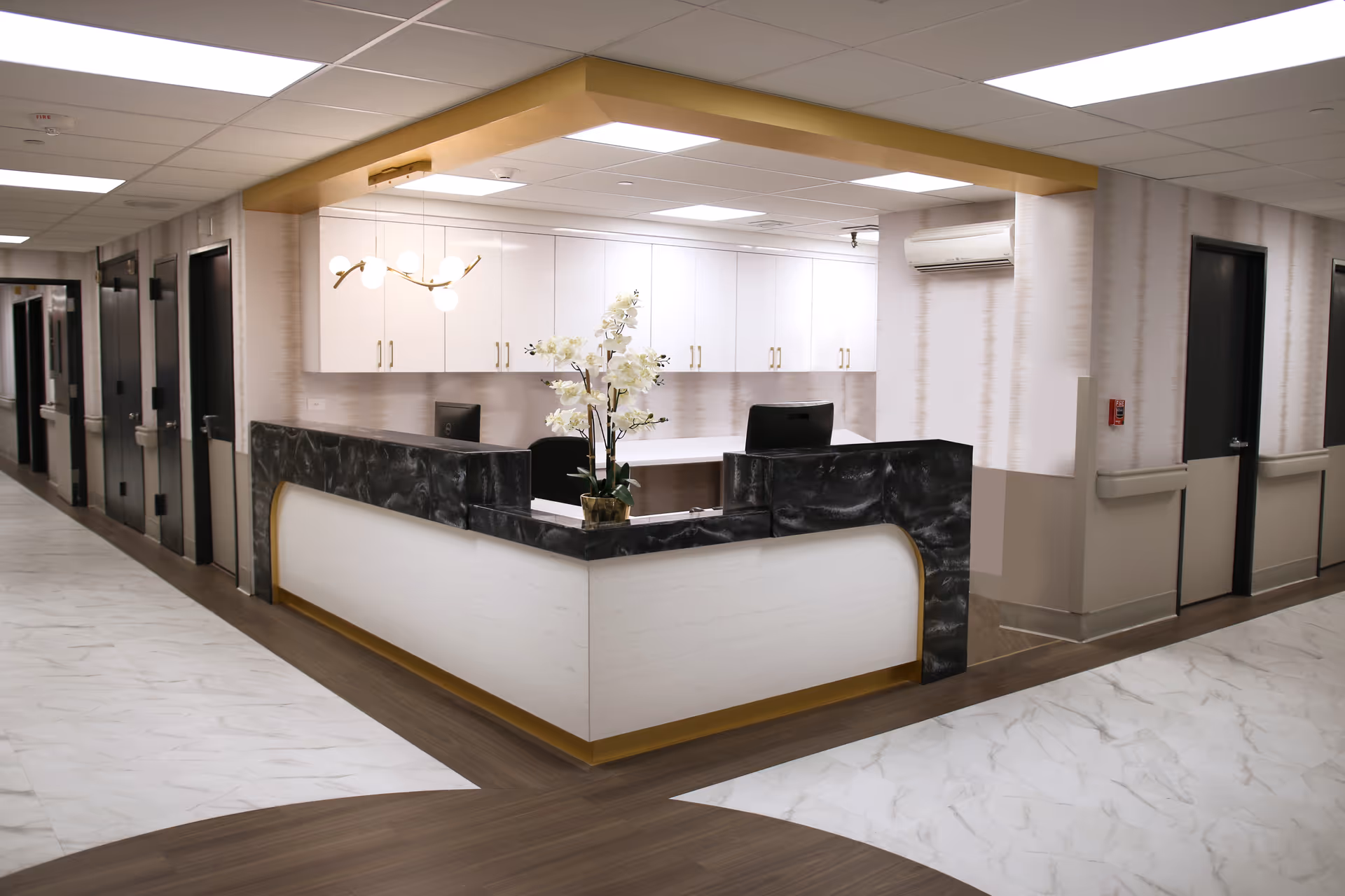 A modern reception desk area in a senior living facility with a black and white marble counter, white cabinets with gold handles, a decorative flower arrangement on the desk, and a hallway with multiple doors leading to rooms.