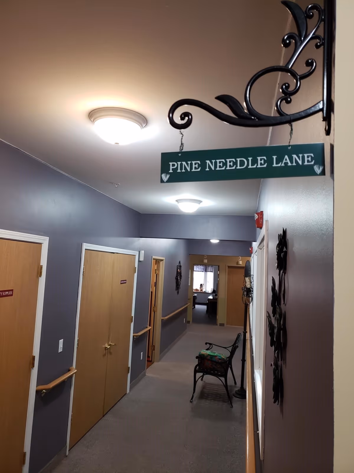 Interior hallway of a senior living facility with gray walls and wooden doors. A decorative hanging sign reads 'PINE NEEDLE LANE'. There is a black metal bench with a cushion and a tall candle holder along the right wall. Handrails run along both sides of the hallway.
