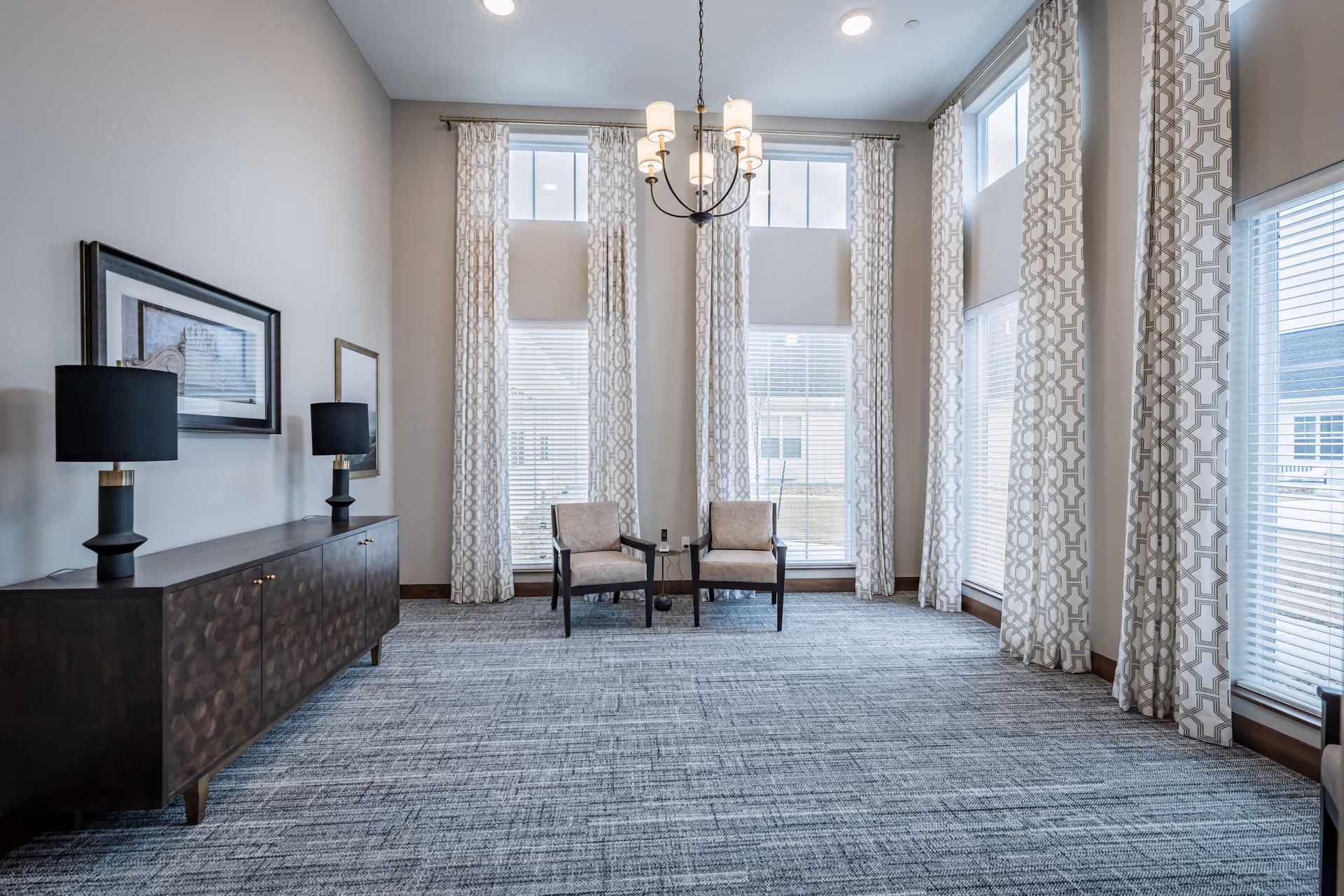 A bright and spacious room with tall windows covered by patterned curtains, two beige armchairs with dark wooden frames positioned around a small table, a dark wooden sideboard with two black lamps, and a chandelier hanging from the ceiling.