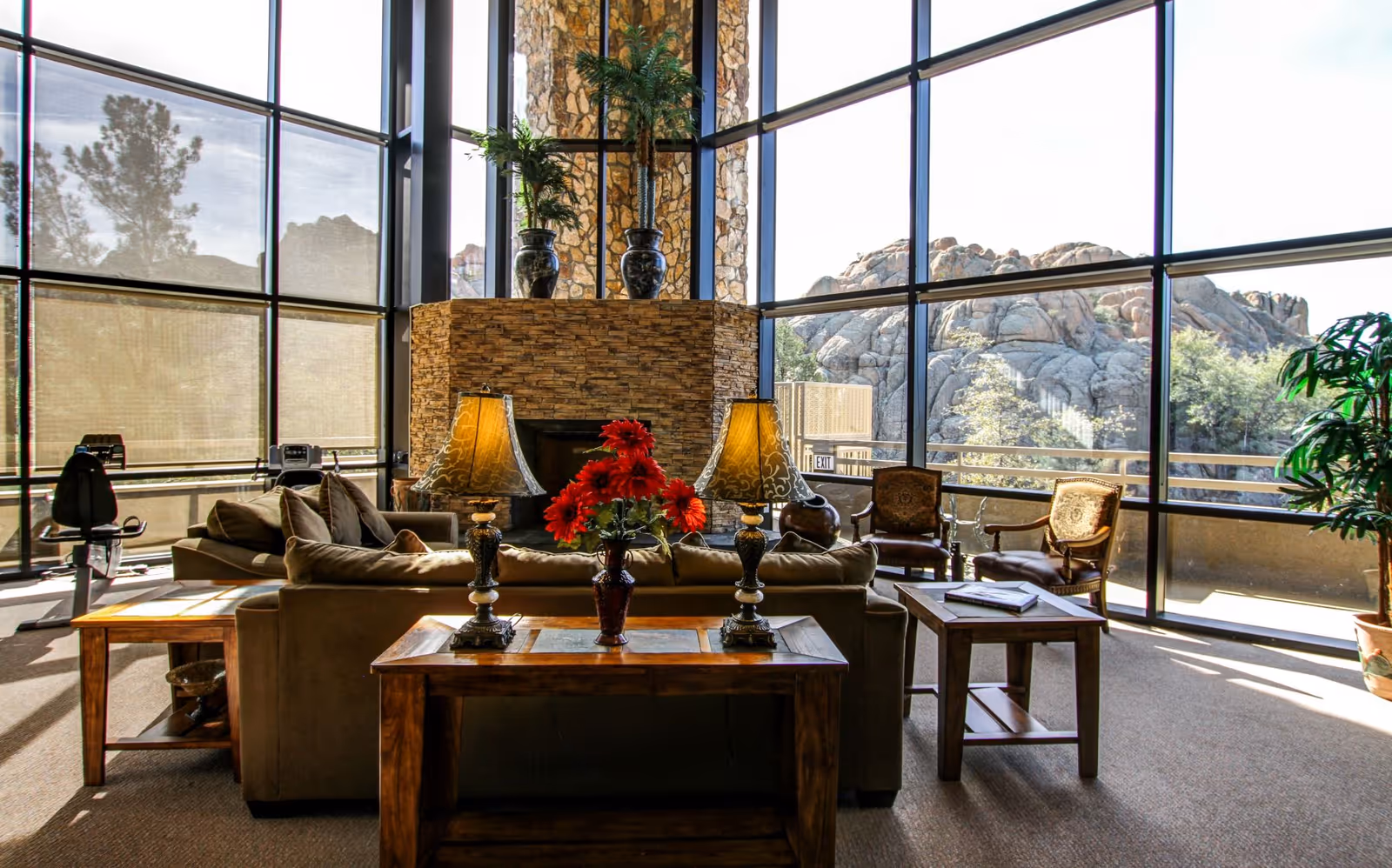 Sunlit common living area with sofas, wooden side tables and lamps in front of a stone fireplace and floor-to-ceiling windows overlooking rocky outcrops.
