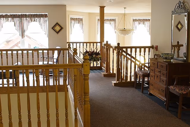 Sunlit upstairs common area with wooden balustrades, seating, a dresser and a decorative mirror.
