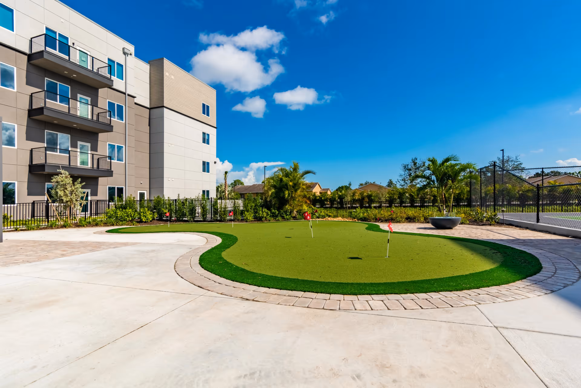 Outdoor area of a senior living facility featuring a small putting green with several red flags, surrounded by a paved walkway. The background shows a multi-story building with balconies, some palm trees, and a clear blue sky with a few clouds.