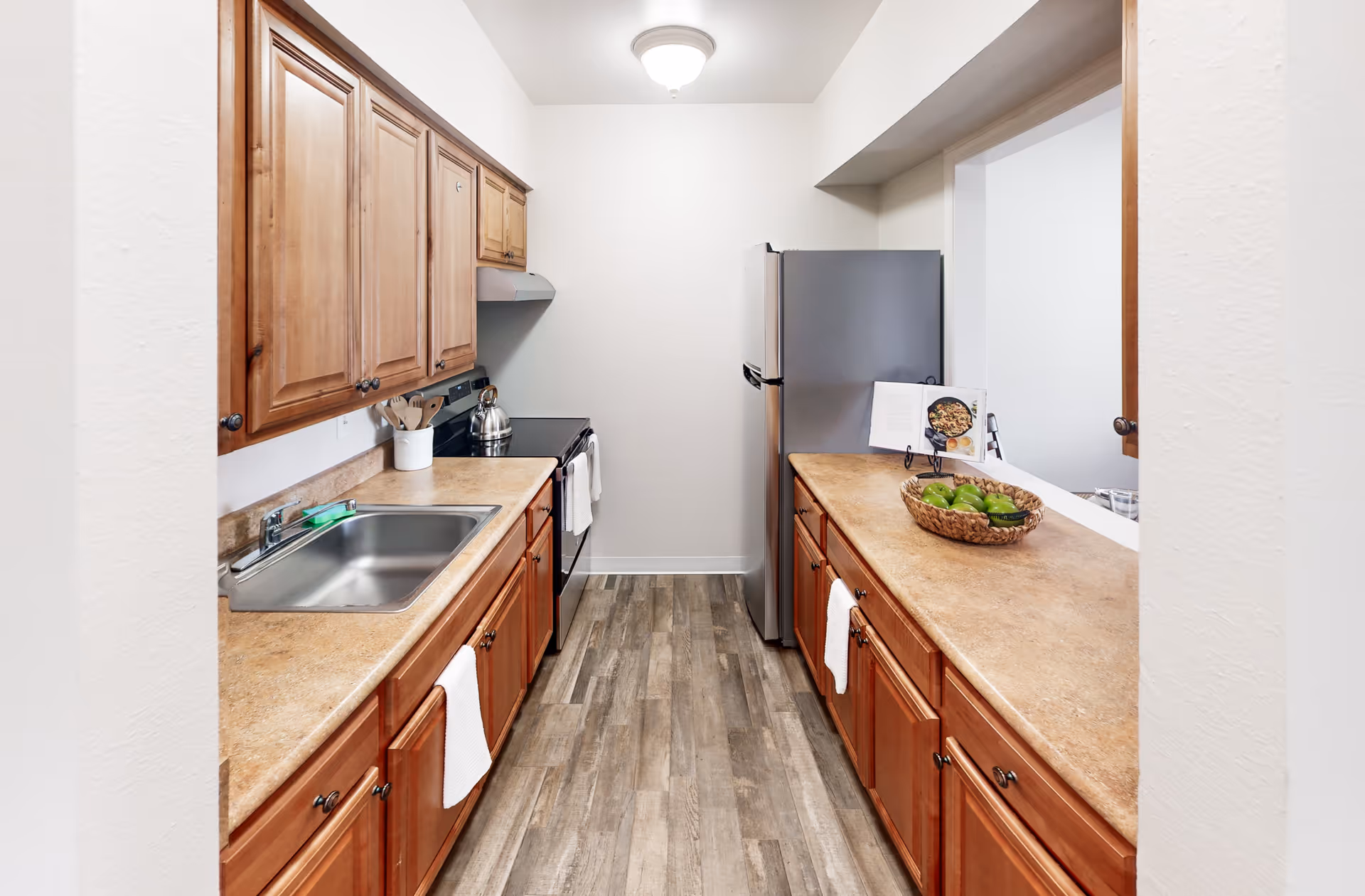A narrow kitchen with wooden cabinets on both sides, beige countertops, a stainless steel sink, a stove with a kettle, and a stainless steel refrigerator at the end. A basket of green apples and a cookbook on a stand are on the right countertop. The floor has wood-like tiles and the walls are white.