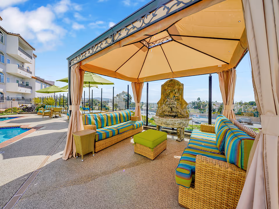 Outdoor seating area at San Clemente Villas featuring a covered gazebo with striped cushioned wicker sofas and a small green ottoman. In the background, there is a decorative stone water fountain, a swimming pool, green umbrellas, and a multi-story building under a blue sky with some clouds.