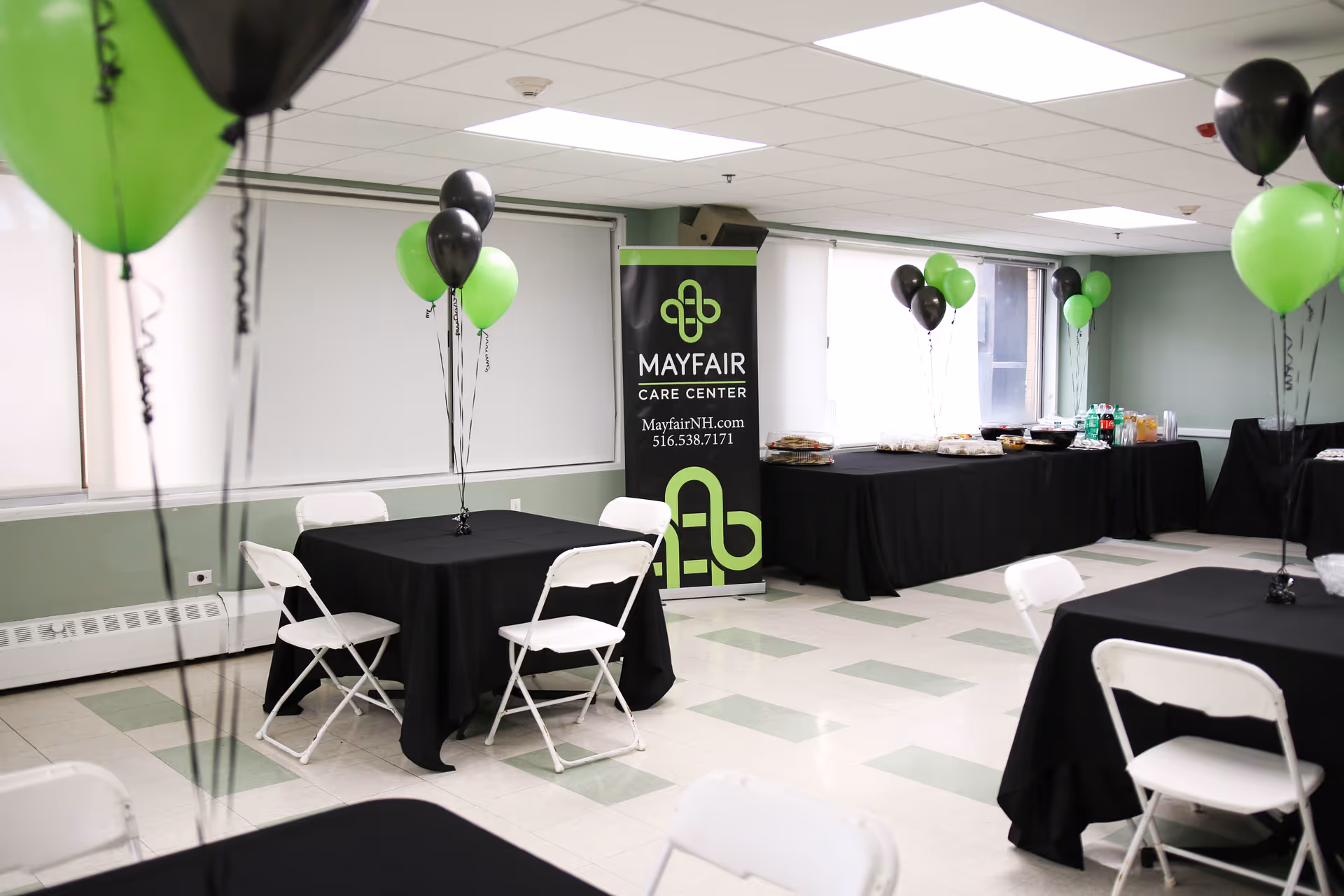 A decorated event room at Mayfair Care Center with black and green balloons tied to tables covered with black tablecloths. White folding chairs surround the tables. A banner with the Mayfair Care Center logo, website, and phone number is displayed near a window. A buffet table with food and drinks is set up along the wall.