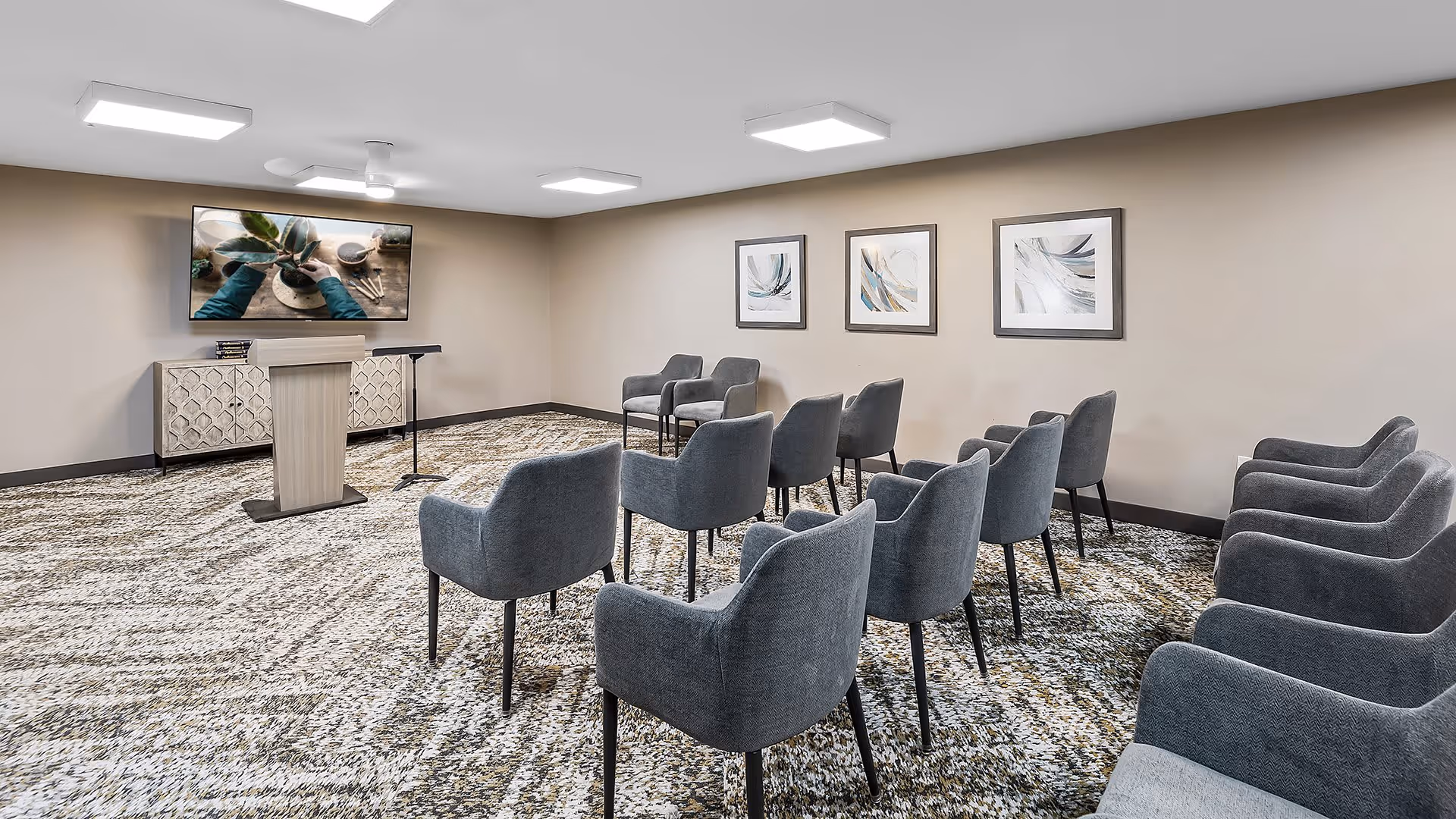 A small conference or presentation room with several gray upholstered chairs arranged in rows facing a podium and a large flat-screen TV mounted on the wall. The room has beige walls with three abstract framed artworks and a patterned carpeted floor. The lighting is provided by ceiling-mounted square lights.