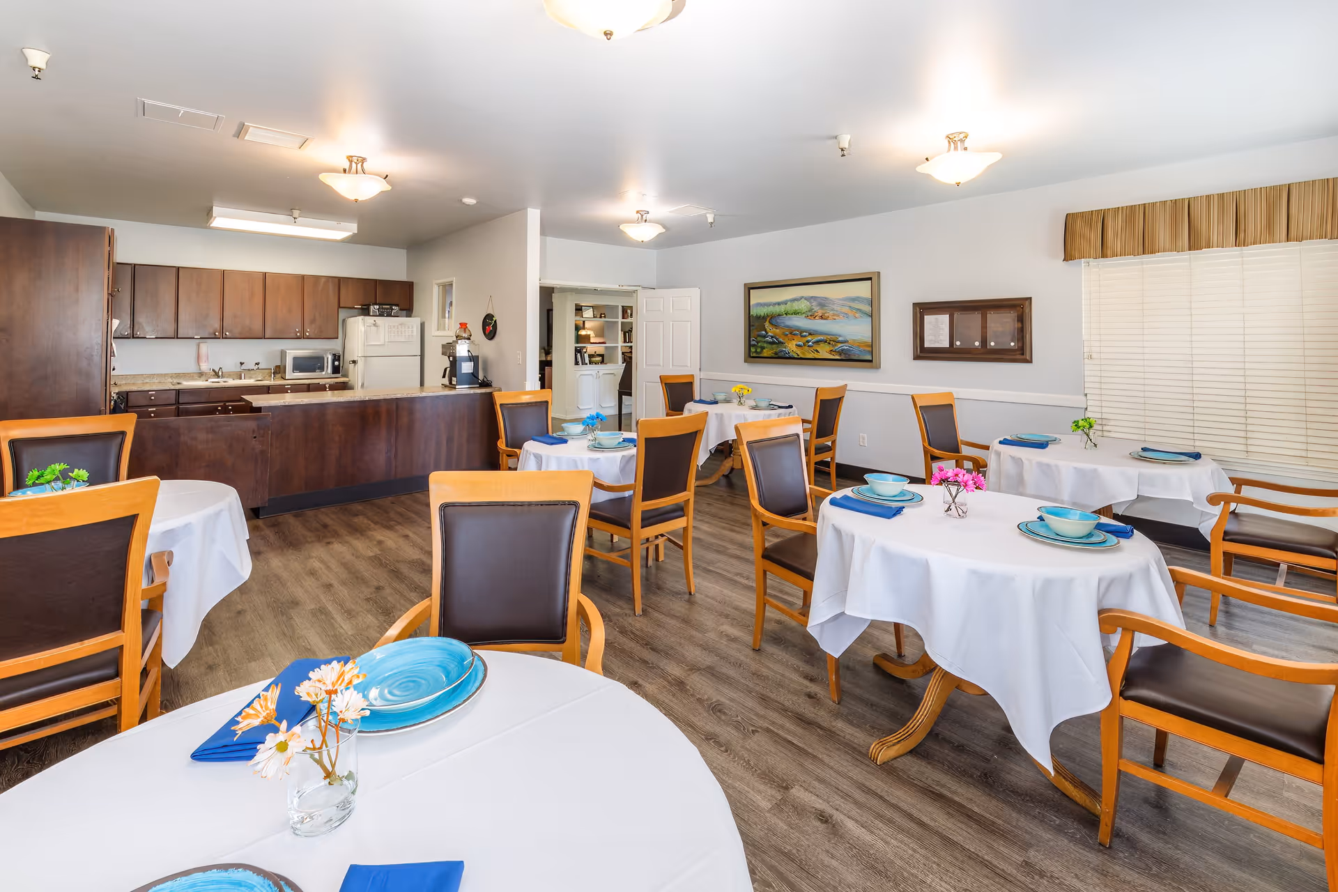 A dining room in an assisted living facility with several round tables covered with white tablecloths, each set with blue plates, bowls, and napkins. Wooden chairs with dark cushions surround the tables. In the background, there is a kitchen area with dark wooden cabinets, a refrigerator, microwave, and coffee maker. The room has wood flooring, white walls, and ceiling lights. A painting and a bulletin board are mounted on the wall, and a window with closed blinds is visible.