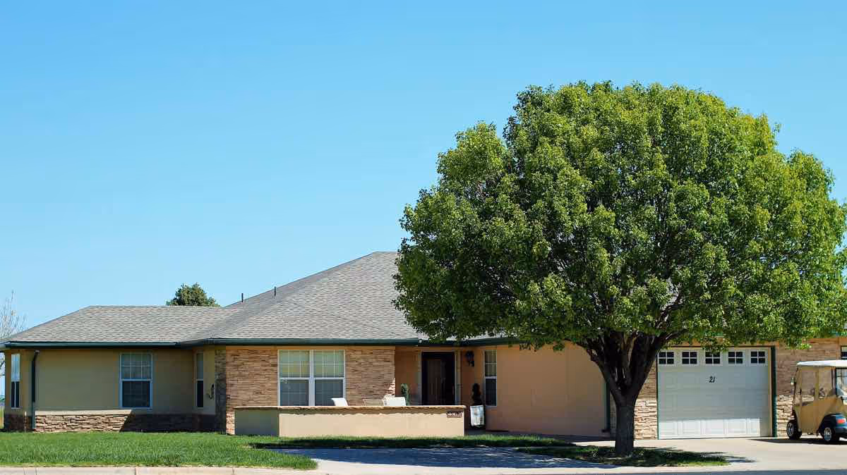 Exterior view of a single-story senior living facility building with a large green tree in front, a driveway, and a golf cart parked near a garage door under a clear blue sky.
