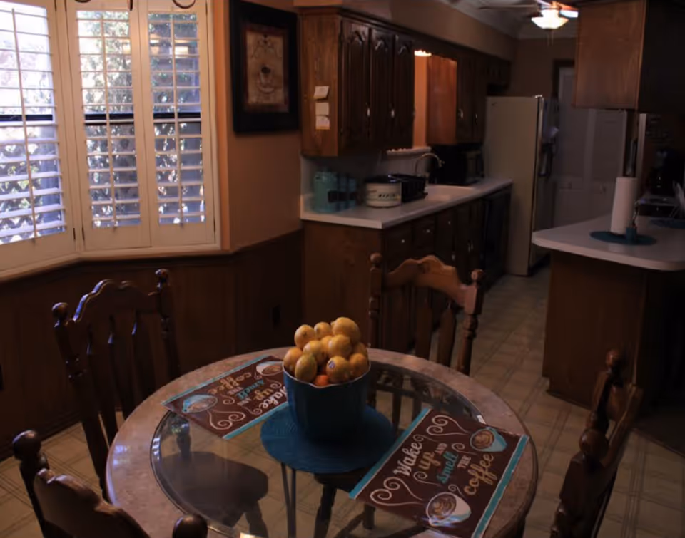 A kitchen dining area with a round glass-top table surrounded by wooden chairs. On the table is a blue bowl filled with fruit and two placemats with coffee-themed designs. The kitchen has wooden cabinets, a white countertop, a refrigerator, and a window with white shutters letting in natural light.