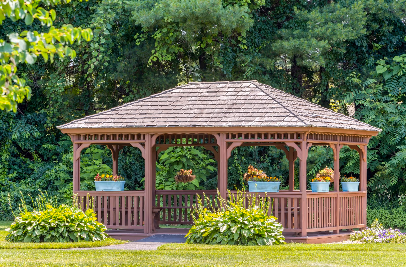 A wooden gazebo with a shingled roof situated in a lush green garden area. The gazebo is surrounded by various plants and flowers, with several flower pots placed on the railing. Tall trees and dense foliage form the background.