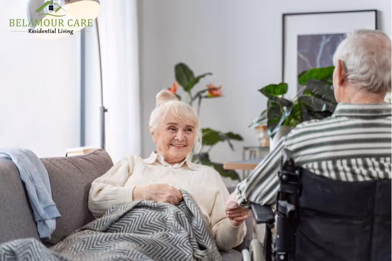 An elderly woman sitting on a couch covered with a blanket, smiling and holding hands with an elderly man in a wheelchair in a cozy living room with plants and framed artwork on the wall.