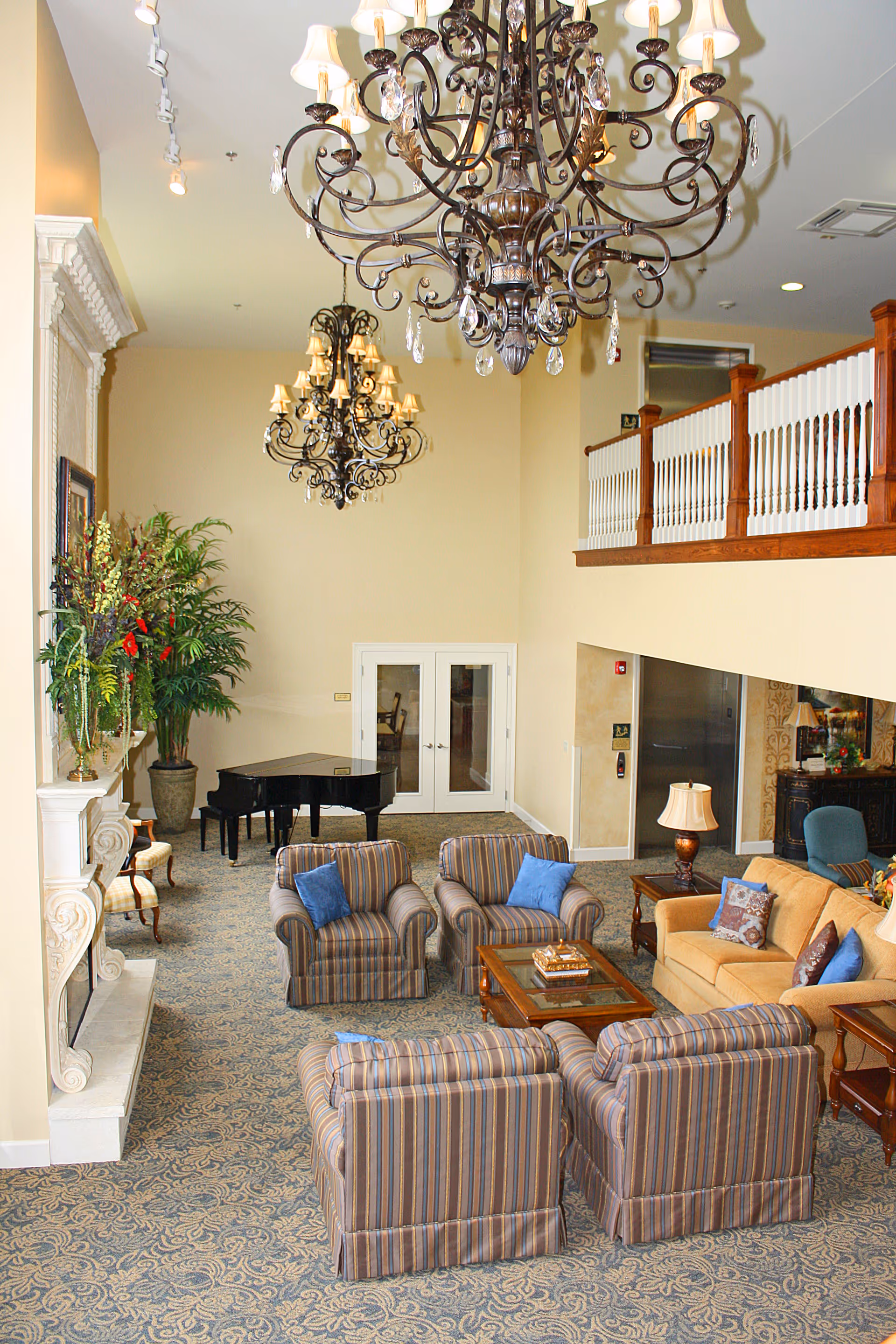 Spacious elegant lounge with chandeliers, grouped upholstered chairs and sofas around a coffee table, a grand piano, fireplace, and an upper balcony.