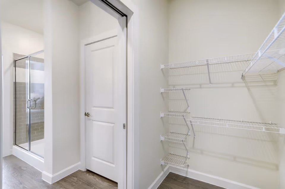 Interior view of a small walk-in closet with white wire shelving on the right wall and a closed white door in the center. To the left, there is a glass-enclosed shower with gray and white tiled walls visible through an open doorway.
