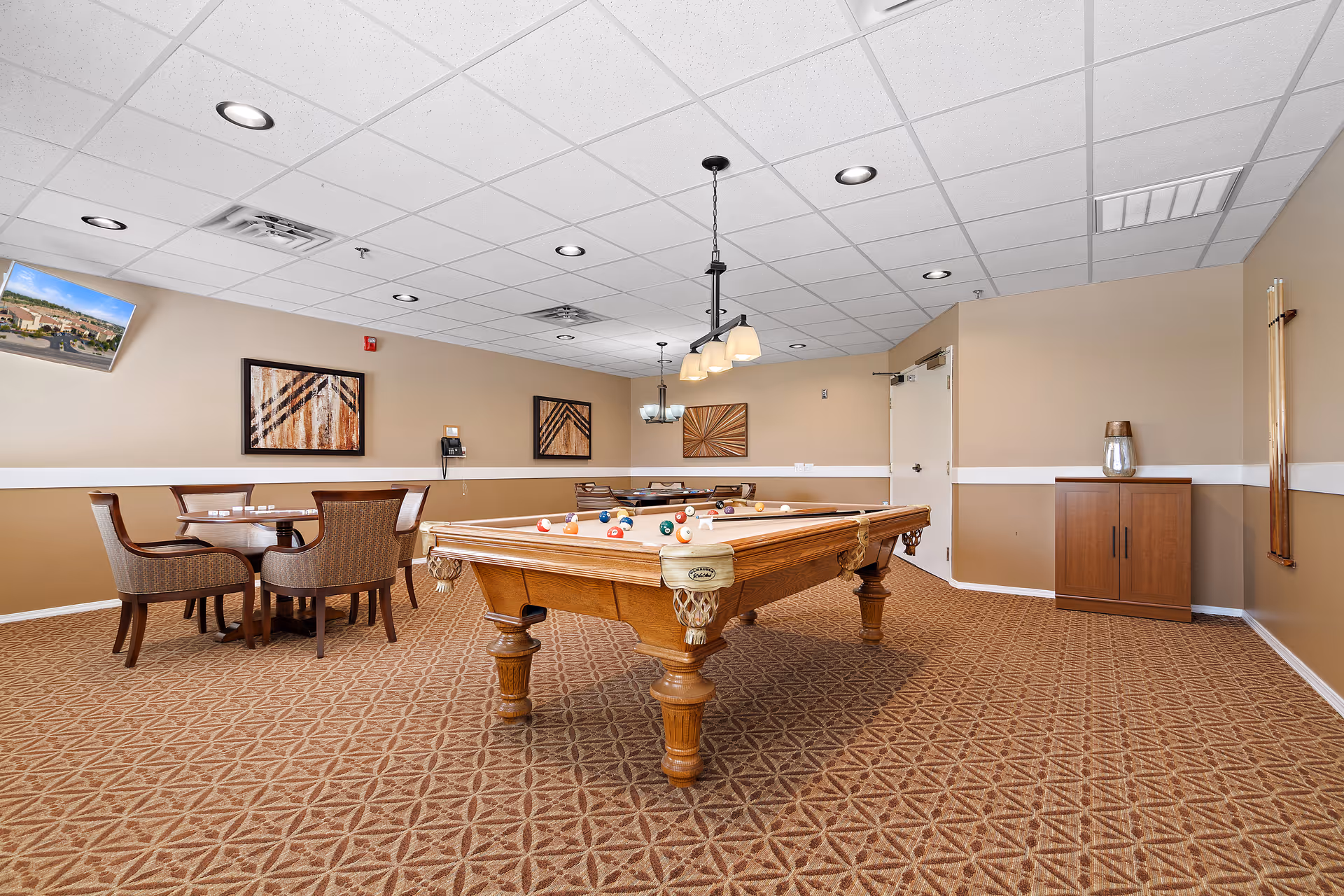 A recreational room with a pool table in the center, a small round table with four chairs in the corner, beige walls with three framed abstract artworks, a wall-mounted TV, a wooden cabinet, and a rack holding pool cues. The room has a patterned carpet and a white drop ceiling with recessed lighting and hanging light fixtures above the pool table.