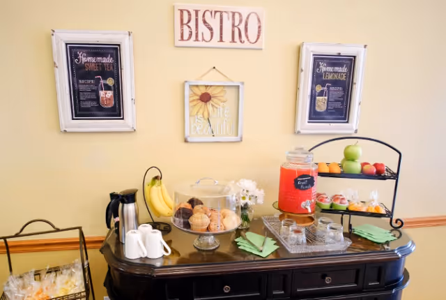 A buffet table set against a beige wall with framed signs reading 'BISTRO', 'Homemade Sweet Tea', 'Homemade Lemonade', and 'Life is beautiful'. The table holds a variety of refreshments including a glass dispenser with fruit punch, muffins and pastries under a glass dome, a basket of bananas, a tiered tray with apples and oranges, a coffee pot, cups, and green napkins.