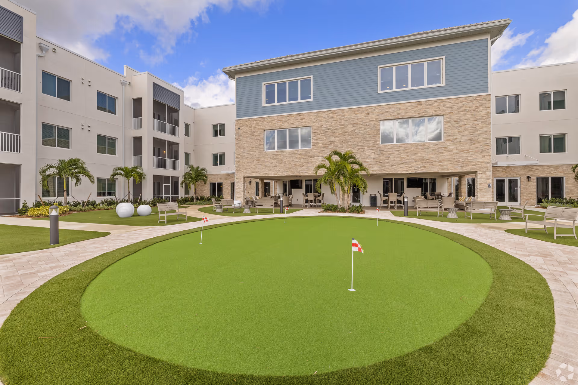 Outdoor courtyard area of a senior living facility featuring a putting green with small flags, surrounded by paved walkways, benches, palm trees, and a three-story building with balconies and large windows under a partly cloudy sky.