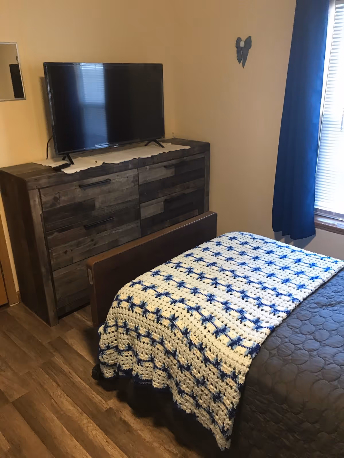 Small bedroom with a bed covered by a blue-and-white crocheted throw, a wooden dresser with a TV, and a window with a blue curtain.