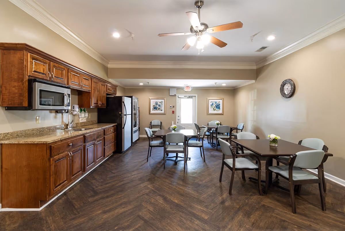 A dining area in a senior living facility featuring several tables with chairs arranged around them. The room has a ceiling fan with lights, wooden flooring, beige walls, and a kitchen area with wooden cabinets, a microwave, a sink, and a refrigerator. There are framed pictures on the far wall and a clock on the right wall.