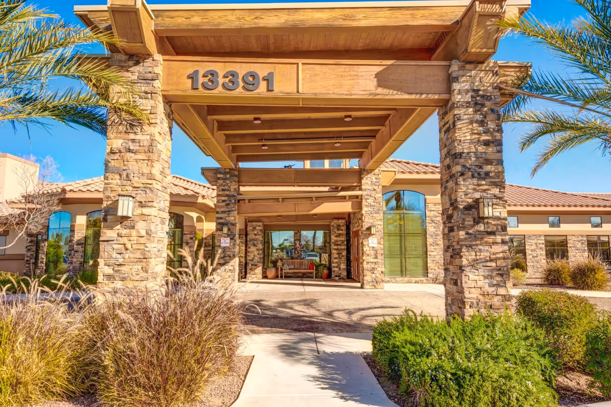 Front entrance of a senior living facility with stone pillars and a wooden canopy displaying the number 13391. The building has large windows and a tiled roof, surrounded by palm trees and landscaping with bushes and grasses under a clear blue sky.