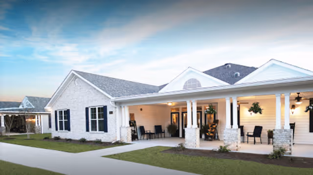 Exterior front of a single-story white brick building with a covered porch, columns, chairs, and a paved walkway under a blue sky.