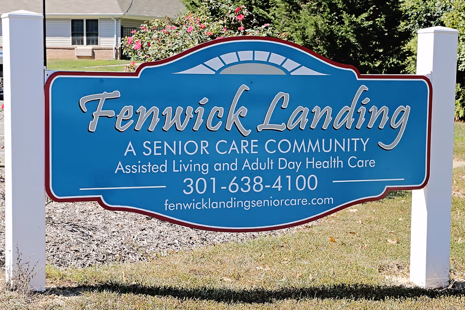 A blue and white outdoor sign for Fenwick Landing Senior Care Community, offering assisted living and adult day health care, with a phone number and website displayed. The sign is mounted on white posts with a background of grass, bushes, and a building.
