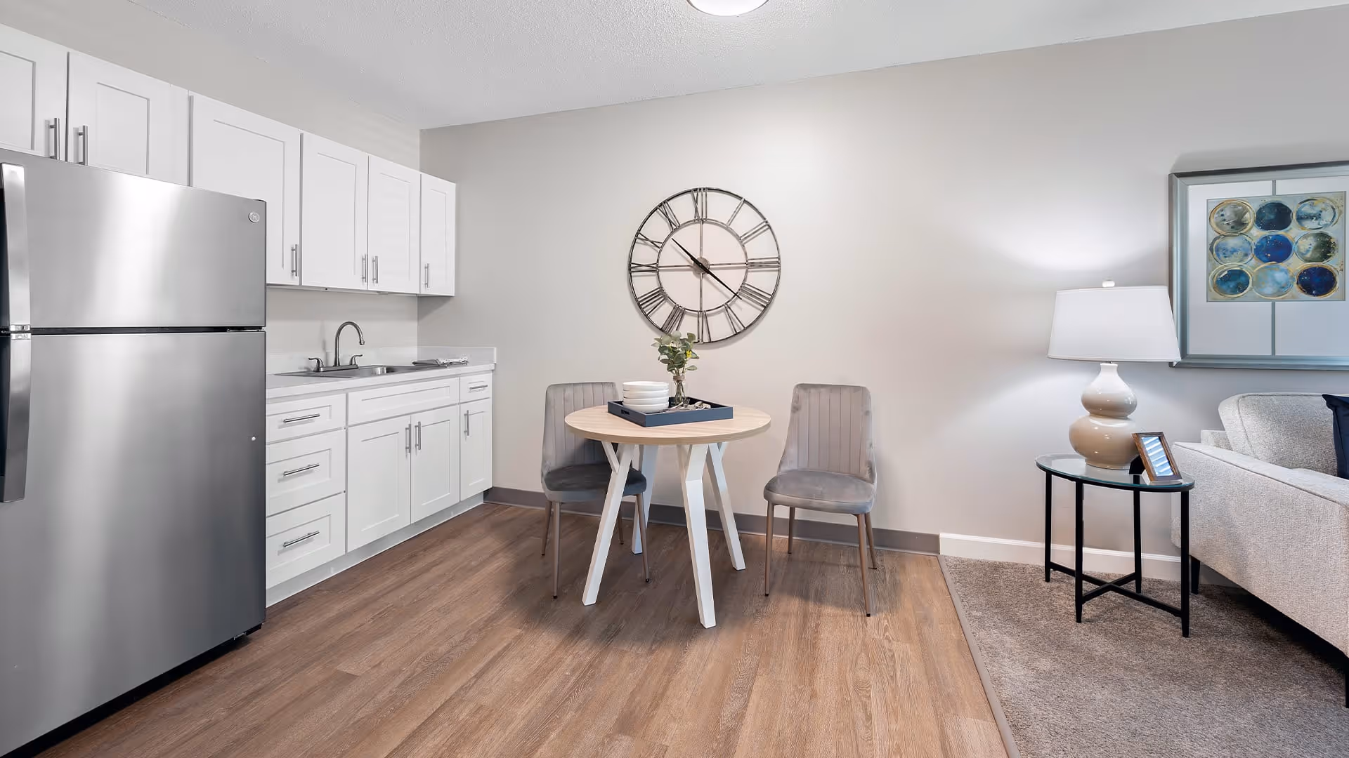 A modern kitchen and small dining area with white cabinets, a stainless steel refrigerator, a round wooden table with two gray chairs, a large wall clock, and a side table with a lamp next to a light-colored sofa.