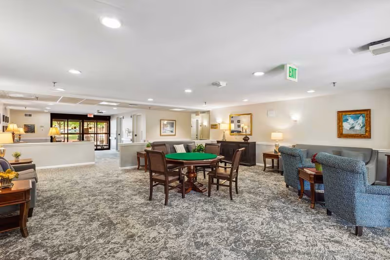 Spacious communal lounge with a central card table, upholstered chairs, side tables, lamps, and framed artwork in a senior living facility.