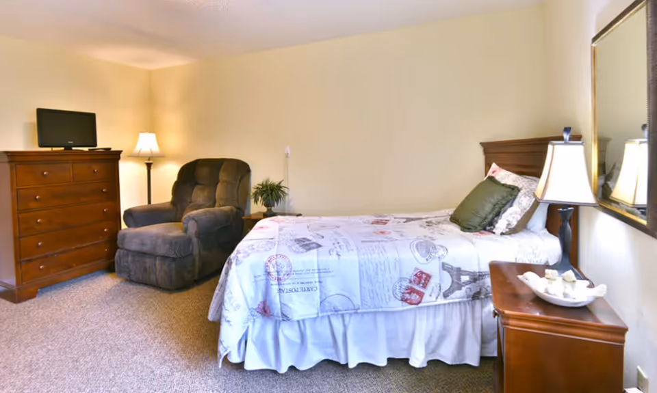 A cozy bedroom with a single bed covered in a white and patterned bedspread, a green pillow, a wooden nightstand with a lamp and decorative items, a large mirror on the wall, a comfortable dark armchair, a wooden dresser with a small TV on top, and a floor lamp in the corner.