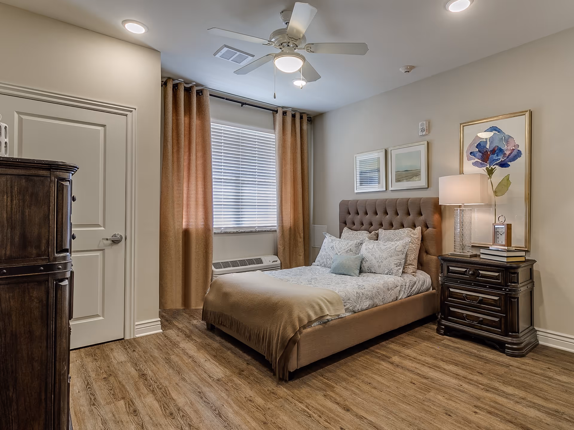 A cozy bedroom with a tufted upholstered bed dressed in patterned bedding and a beige throw blanket. There are three framed artworks on the wall above the bed. To the right of the bed is a dark wooden nightstand with a lamp, a clock, and some books. The room has wood flooring, beige walls, a window with closed blinds and brown curtains, a ceiling fan with a light, and a closed white door on the left side.