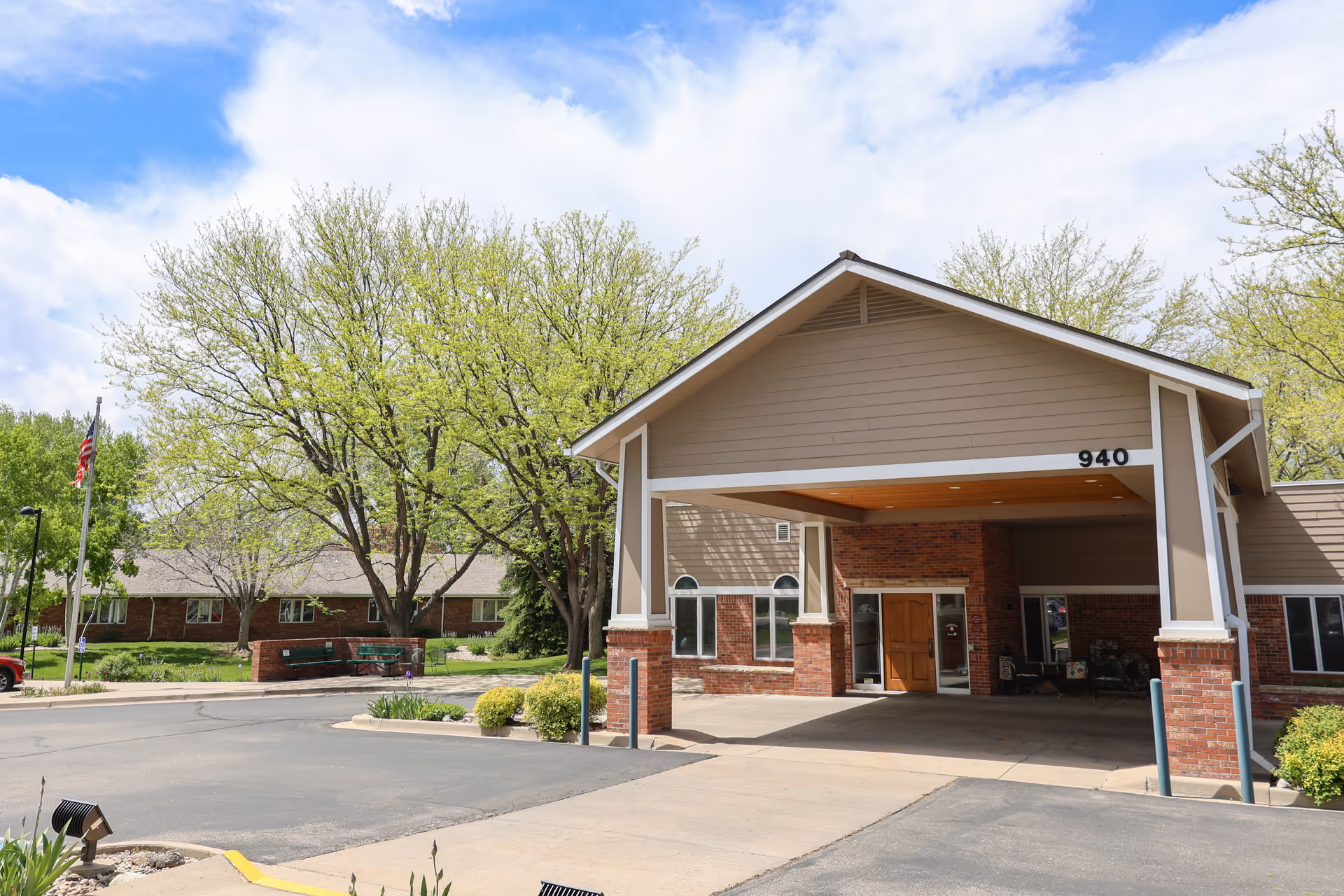 Exterior view of Columbine West Health & Rehab Facility showing the entrance with a covered drop-off area, brick and beige siding, surrounded by trees and landscaping under a partly cloudy sky.