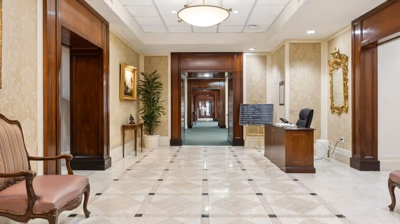 A spacious, well-lit hallway with polished tile flooring and beige patterned wallpaper. The hallway features wooden door frames, a potted plant, a small table with a decorative sculpture, a framed painting, and a wooden desk with a chair and a signboard on an easel. A gold-framed mirror hangs on the right wall.