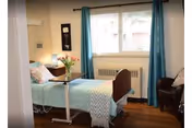 A senior living facility bedroom with a bed, blue curtains, and flowers on a nightstand.