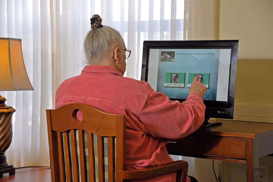 An elderly woman with gray hair tied in a bun, wearing a red jacket, sits on a wooden chair at a wooden desk. She is interacting with a touchscreen computer displaying a quiz question about Winthrop M. Crane. A lamp with a beige shade is on a table to the left, and sheer curtains cover the window in the background.