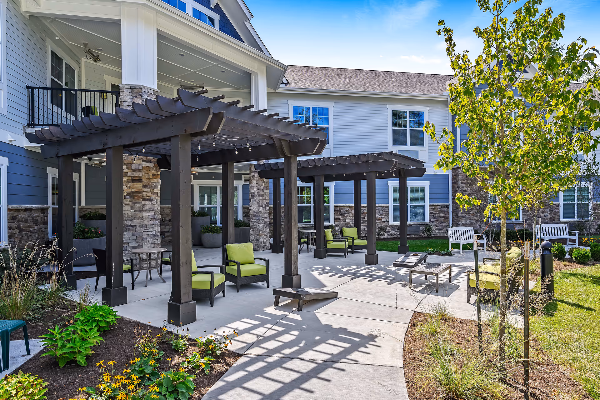 Outdoor patio area at a senior living facility with two dark wooden pergolas, green cushioned chairs, small tables, and landscaped garden beds with flowers and small trees. The building exterior features stone and blue siding with multiple windows under a clear blue sky.